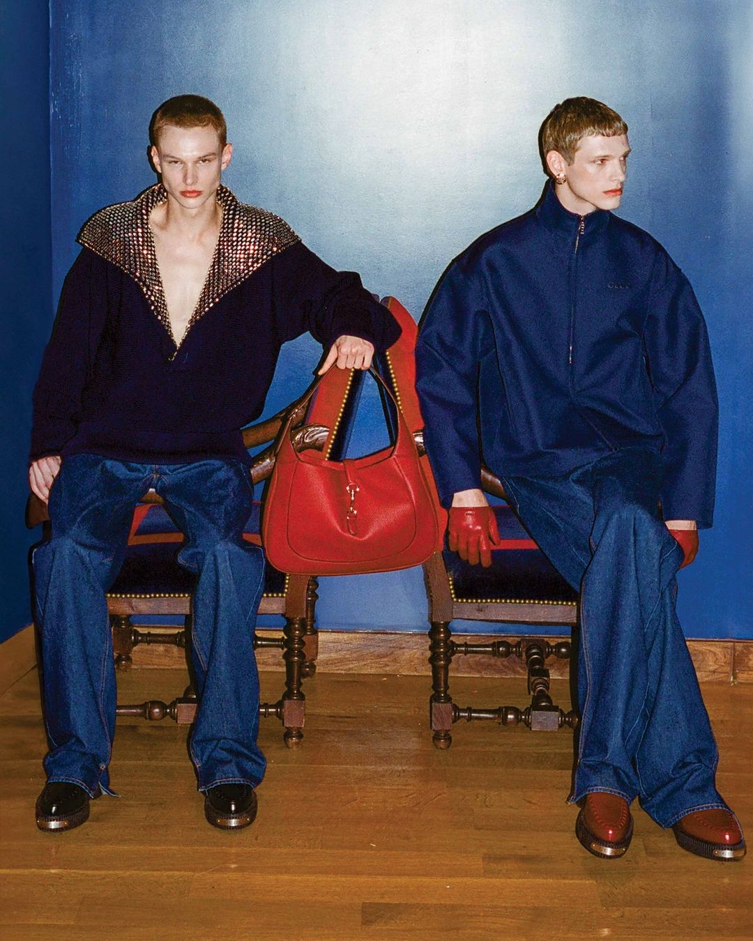 Two young adult fashion models sit on chairs against a blue wall, one holding a red bag, styled in contemporary clothing.