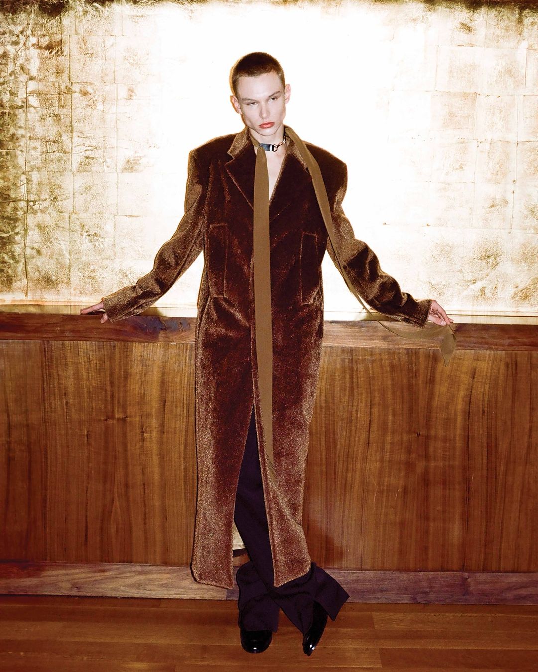 A person in a long brown velvet coat with a brown scarf, posing in front of a gold and wood paneled wall in a portrait photograph.