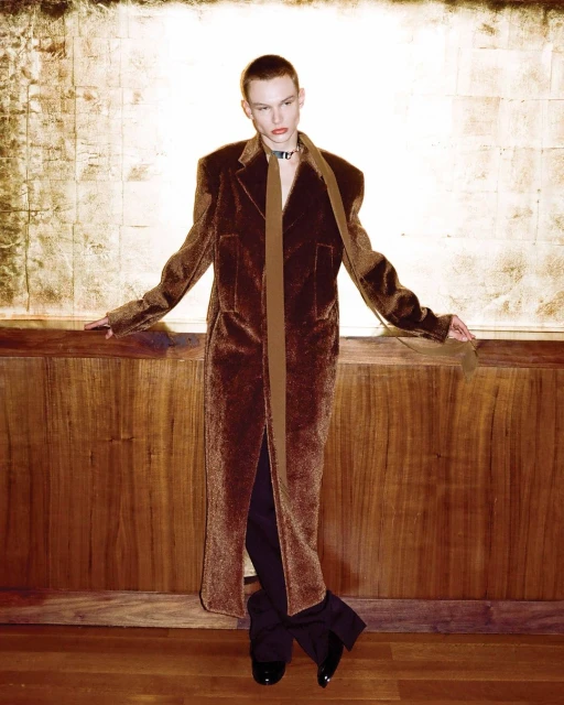 A person in a long brown velvet coat with a brown scarf, posing in front of a gold and wood paneled wall in a portrait photograph.