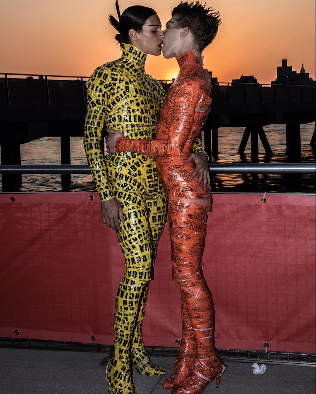 Two people in matching form-fitting outfits kiss on a pier against a sunset. One is yellow with black logos, the other is orange.