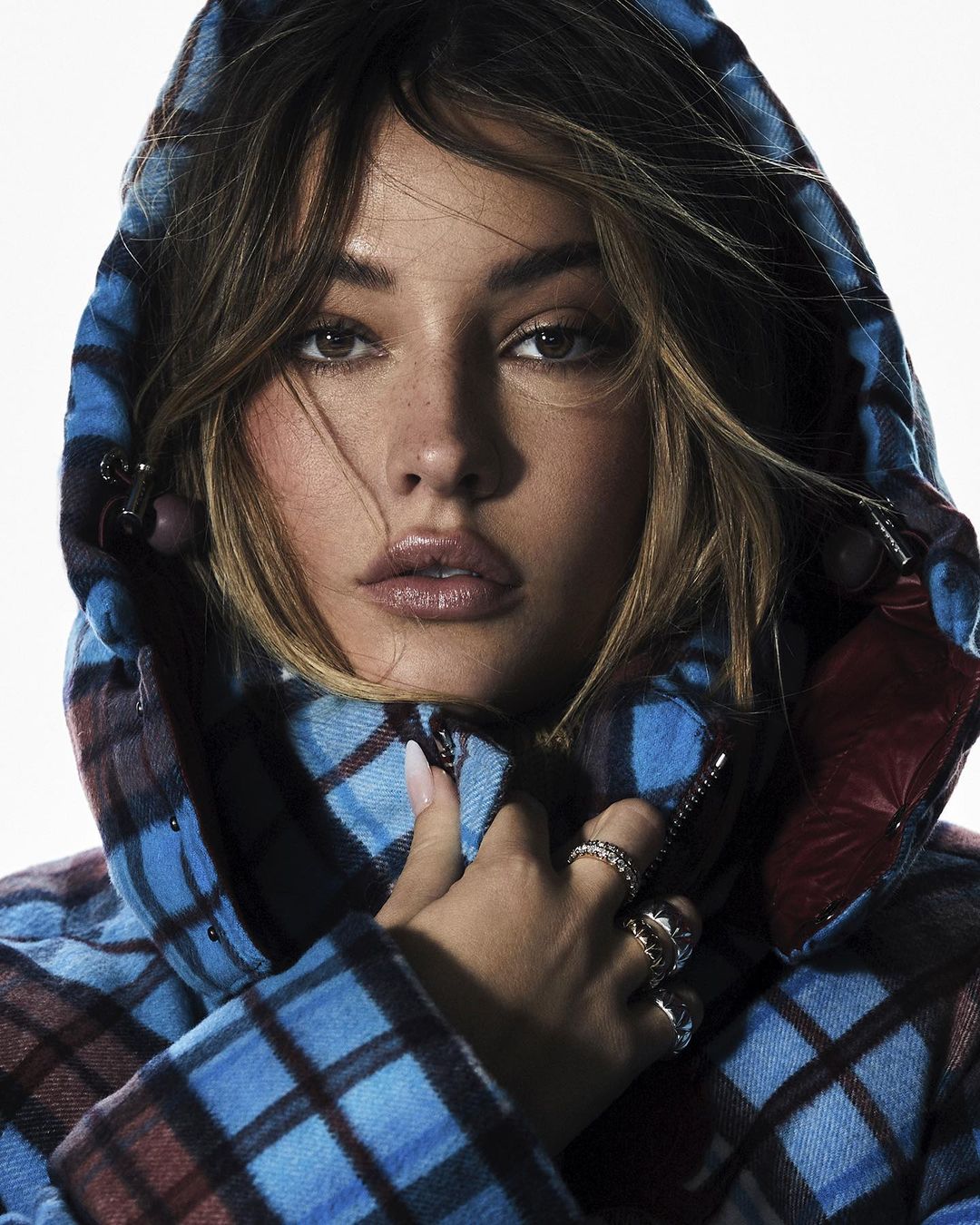 Close-up portrait of a woman with light brown hair and fair skin, wearing a blue and red plaid puffer jacket with the hood up.