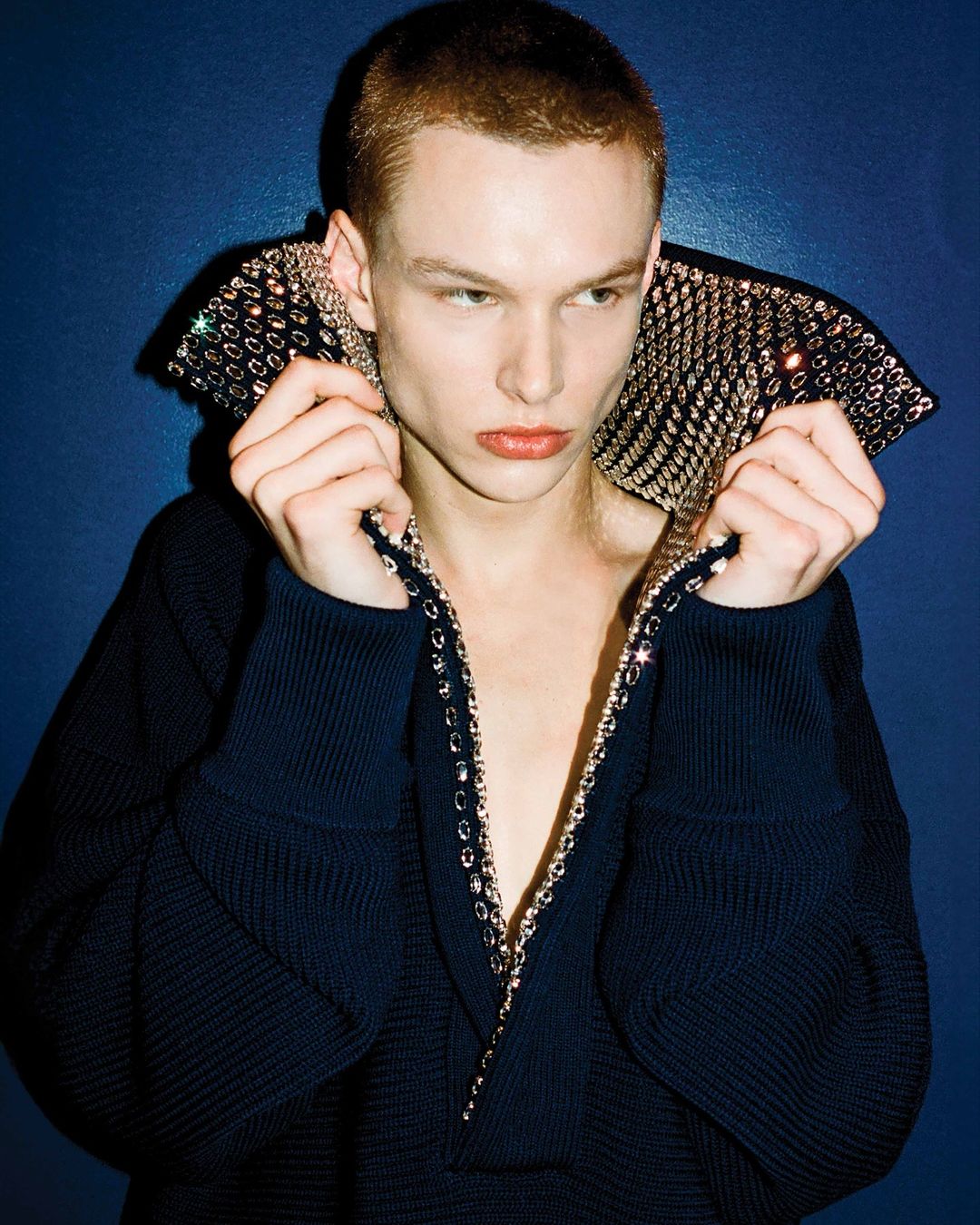 A young adult with short hair in a dark sweater with metallic trim and a large collar poses in front of a dark blue backdrop.