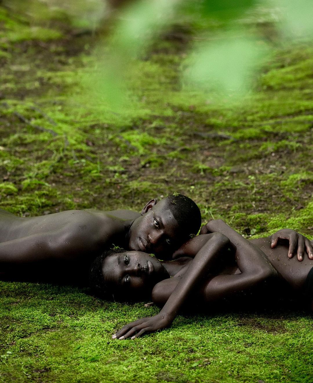 Two dark-skinned adults lie together on moss-covered ground in a close-up, eye-level portrait showcasing natural outdoor beauty and intimate connection.