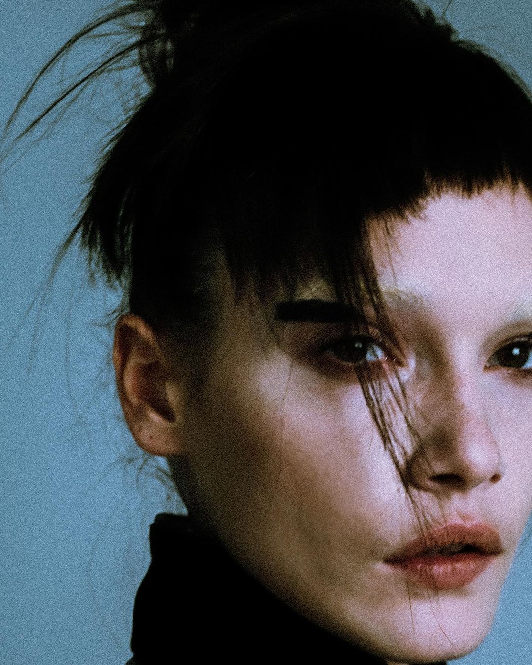 Close-up portrait of an adult female with a dark, messy topknot, short bangs, and neutral expression, set against a light blue background.