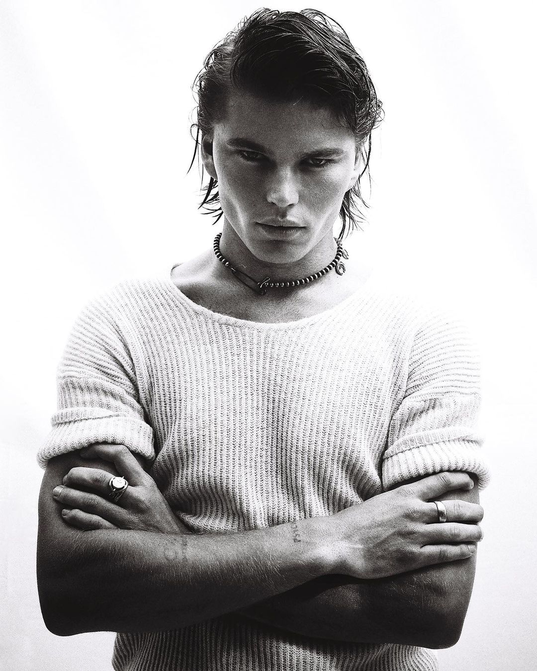 Portrait of a handsome man with dark hair wearing a knit sweater and necklace, standing in front of a white background.