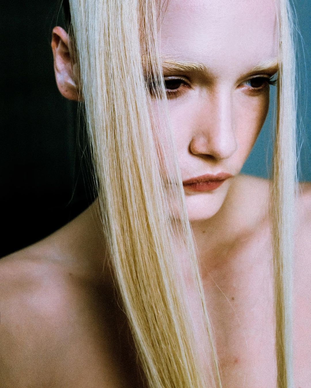 Close-up studio portrait of a young adult woman with pale skin and blonde hair looking down, styled in a beauty fashion shot.