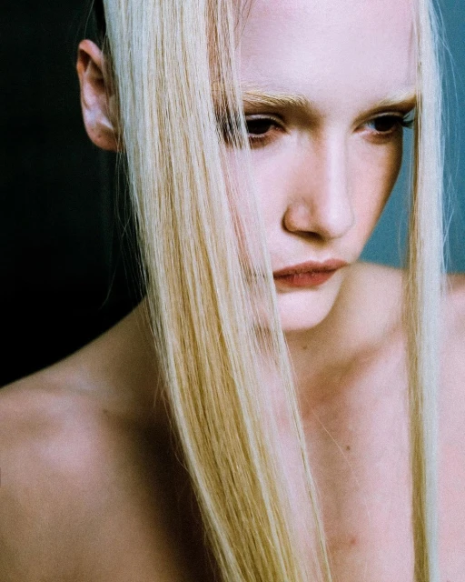 Close-up studio portrait of a young adult woman with pale skin and blonde hair looking down, styled in a beauty fashion shot.