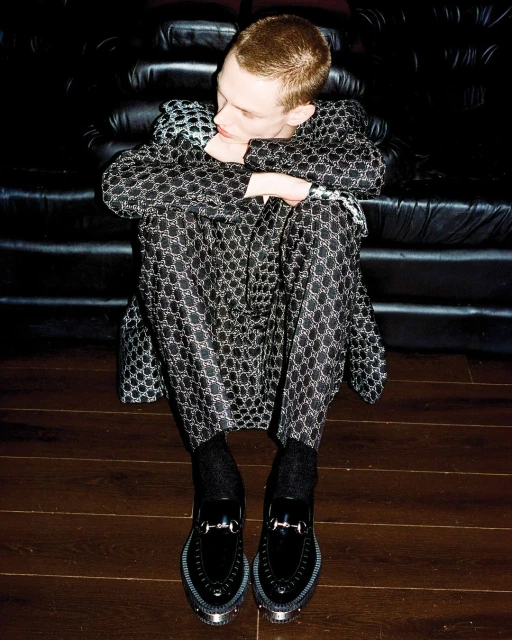 Young adult poses in patterned suit with patent leather loafers indoors. Fashion editorial shot with a relaxed pose.