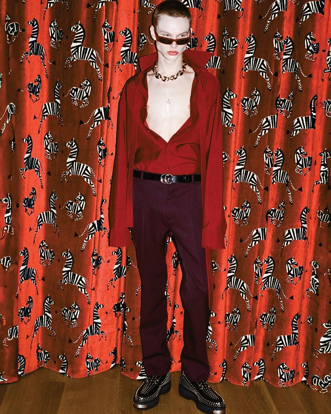 A young, slender man with red hair poses in a red collared shirt, purple pants, and black shoes in front of a zebra-print curtain.