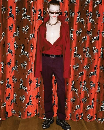 A young, slender man with red hair poses in a red collared shirt, purple pants, and black shoes in front of a zebra-print curtain.