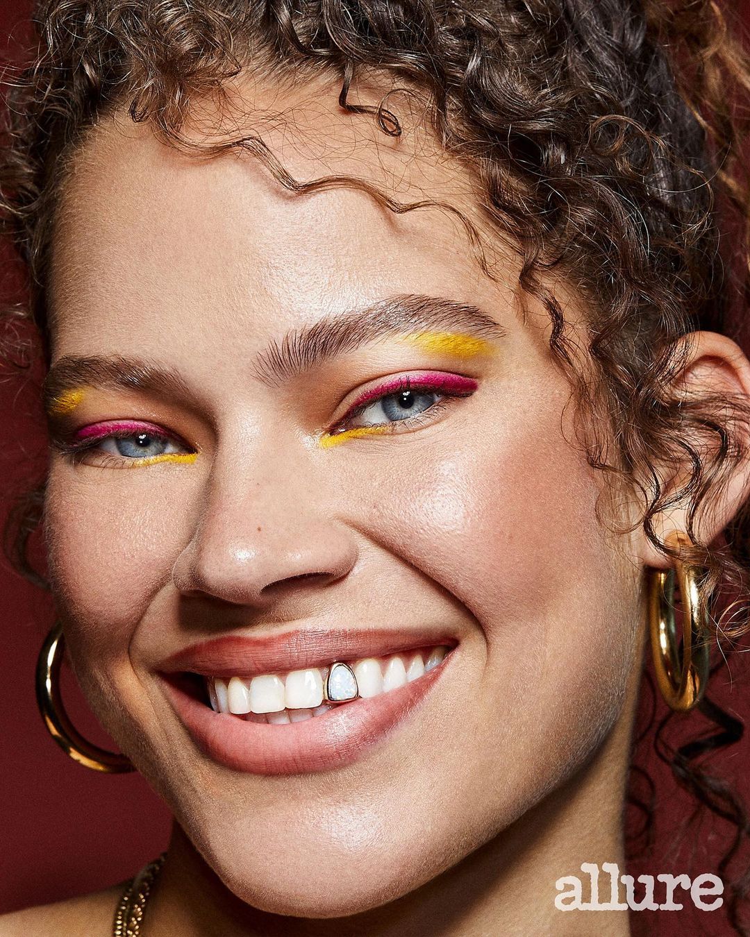 Close-up portrait of a smiling adult female with curly hair, wearing yellow and pink eyeshadow, gold jewelry, and a tooth gem.