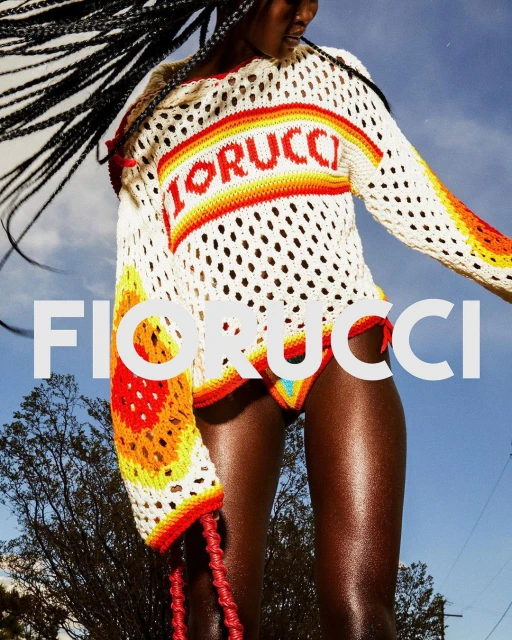 Fashion shot of a dark-skinned model wearing a white knit FIORUCCI top and bikini bottoms outdoors against a blue sky and tree backdrop.
