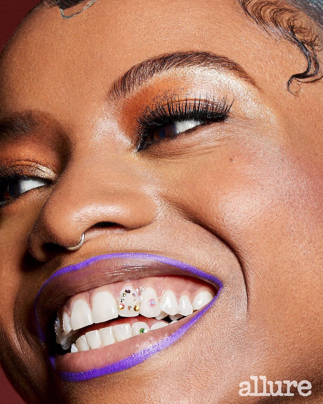 Close-up shot of a smiling woman with purple eyeliner and jeweled teeth. The 'allure' logo is in the bottom right corner.