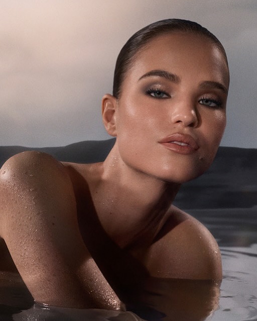 Close-up of a woman partially submerged in water, showcasing her face, shoulders, and wet skin with water droplets. She has a neutral expression.