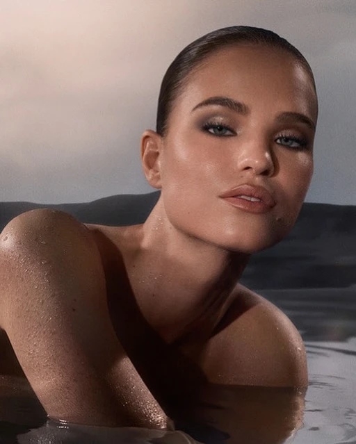 Close-up of a woman partially submerged in water, showcasing her face, shoulders, and wet skin with water droplets. She has a neutral expression.