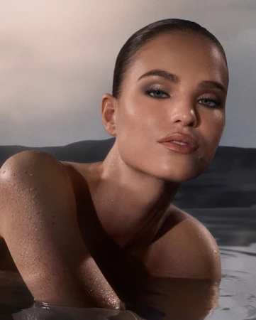 Close-up of a woman partially submerged in water, showcasing her face, shoulders, and wet skin with water droplets. She has a neutral expression.