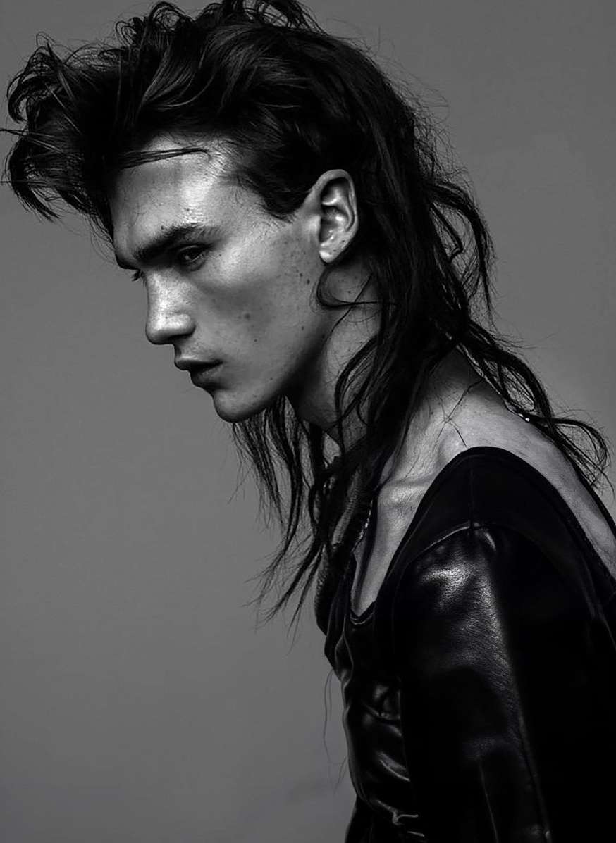 Portrait of a brunette male fashion model with long hair and a leather jacket posing in a photo studio against a gray background.