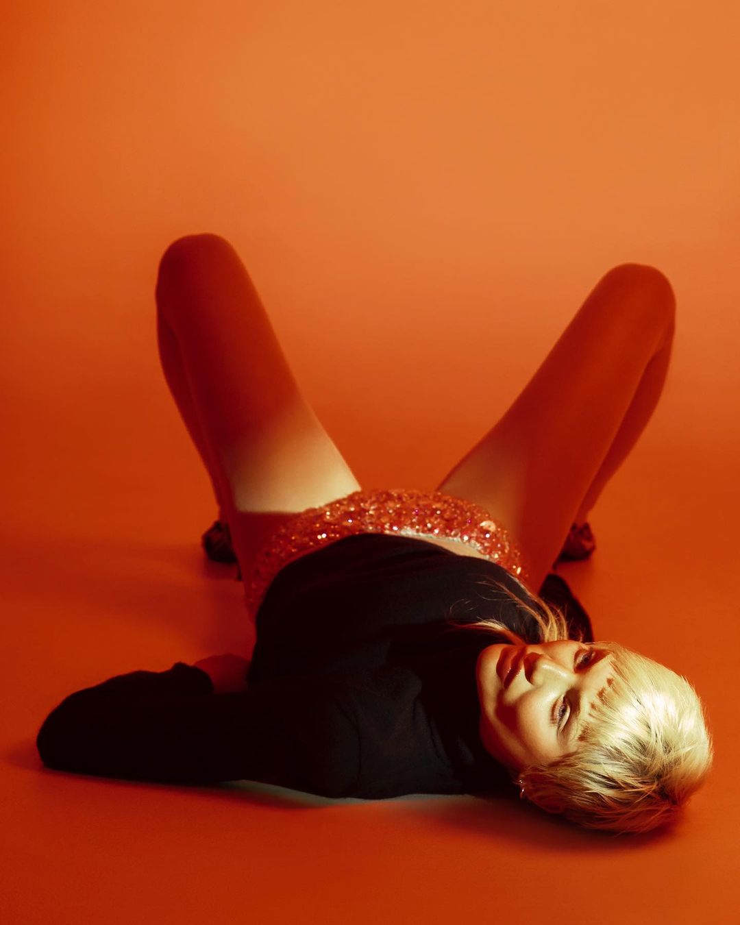 Woman with blonde hair lying on an orange surface wearing a black long-sleeved shirt and sequined skirt, shot in a studio with bright lighting.