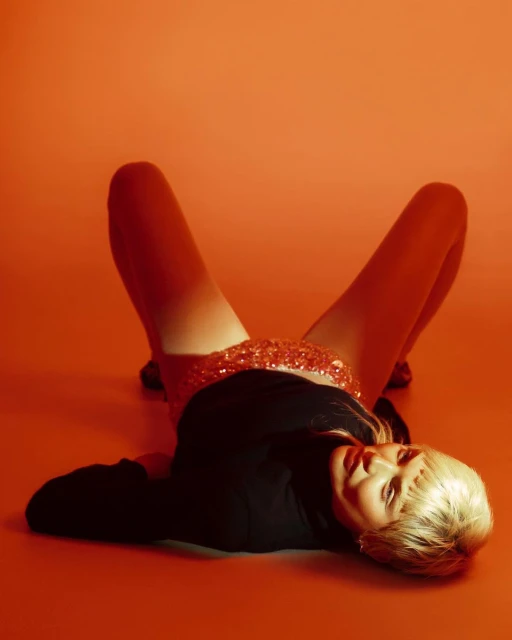 Woman with blonde hair lying on an orange surface wearing a black long-sleeved shirt and sequined skirt, shot in a studio with bright lighting.