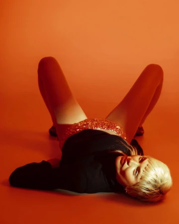 Woman with blonde hair lying on an orange surface wearing a black long-sleeved shirt and sequined skirt, shot in a studio with bright lighting.