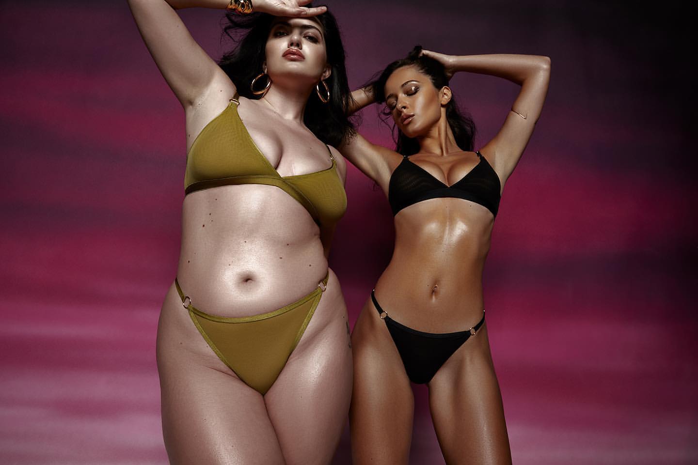Two women pose in bikinis against a pink backdrop, one in olive green and the other in black, accessorized with hoop earrings.