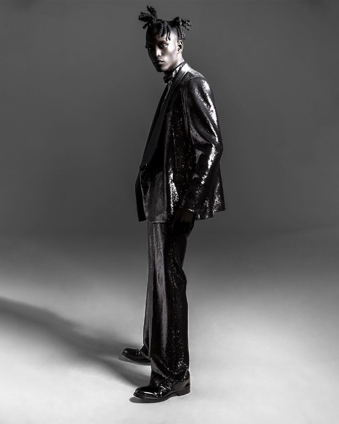 Full-length studio shot of a male fashion model in a black sequined suit looking at the camera with a neutral facial expression.