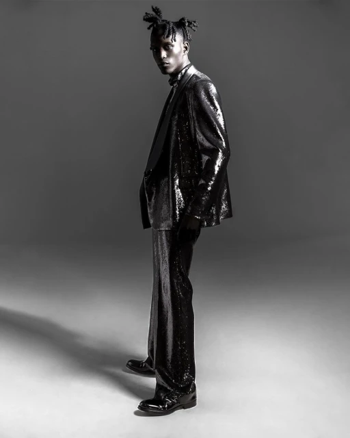 Full-length studio shot of a male fashion model in a black sequined suit looking at the camera with a neutral facial expression.