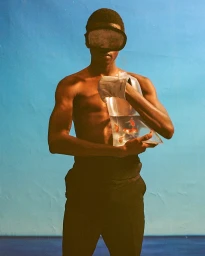 Shirtless man wearing swimming goggles and holding a plastic bag containing goldfish against a bright blue background.