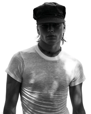 Monochrome portrait of a man wearing a dark flat cap and white t-shirt against a bright white background.