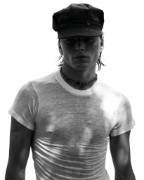 Monochrome portrait of a man wearing a dark flat cap and white t-shirt against a bright white background.