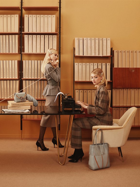 Two women in vintage office setting, one on the phone, one typing on a typewriter, wearing vintage suits with matching handbag and shoes.