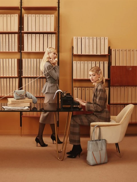 Two women in vintage office setting, one on the phone, one typing on a typewriter, wearing vintage suits with matching handbag and shoes.