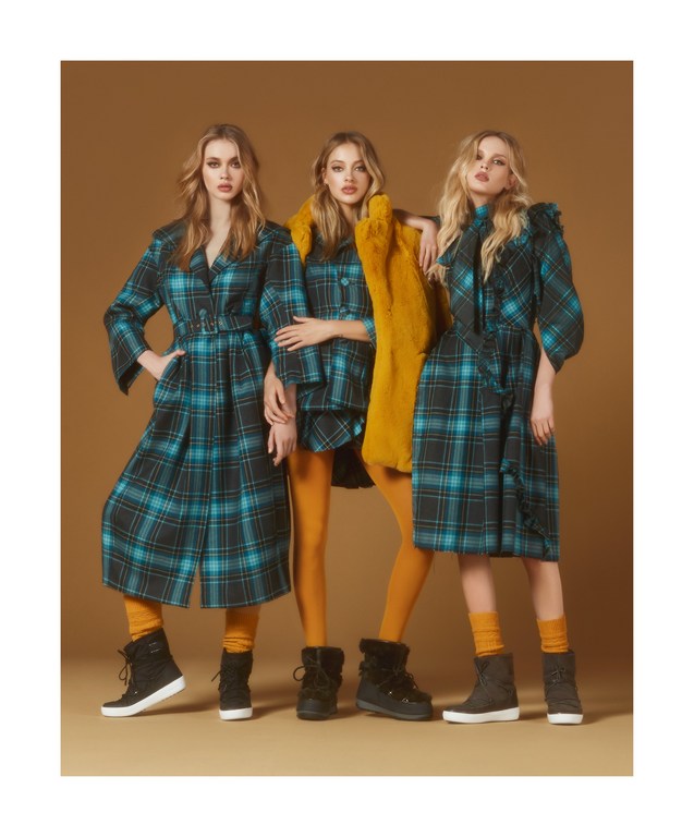 Three fashion models pose in plaid coats and yellow tights against a warm brown backdrop for a studio fashion editorial.