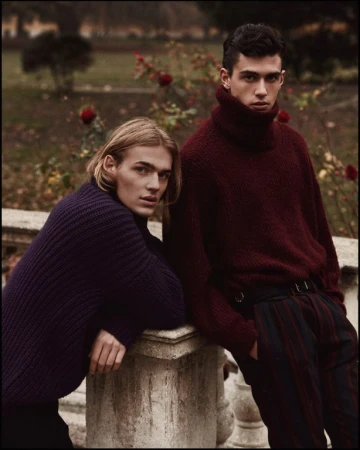 Two young male models leaning against a stone balustrade in a park wearing thick knit turtleneck sweaters in burgundy and navy.
