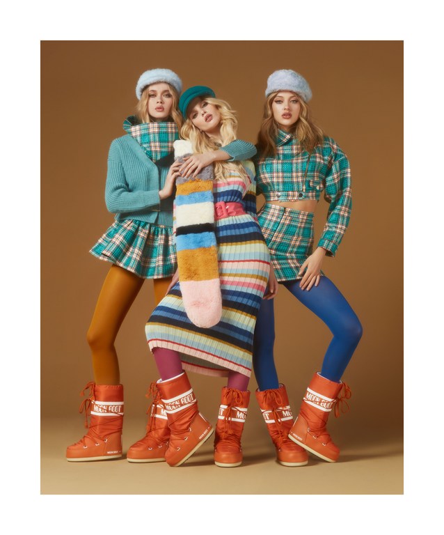Three fashion models pose in a photo studio wearing colorful winter apparel including hats, scarves, dresses, leggings and moon boots.