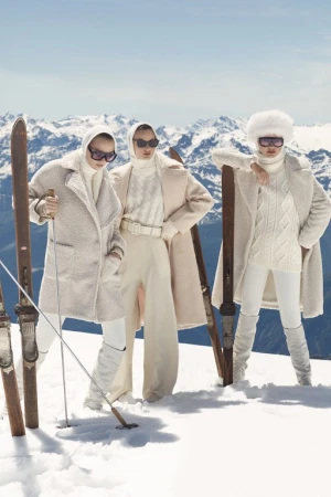 Three women in fashionable winter clothing stand on a snow-covered mountain with vintage skis against a bright blue sky.