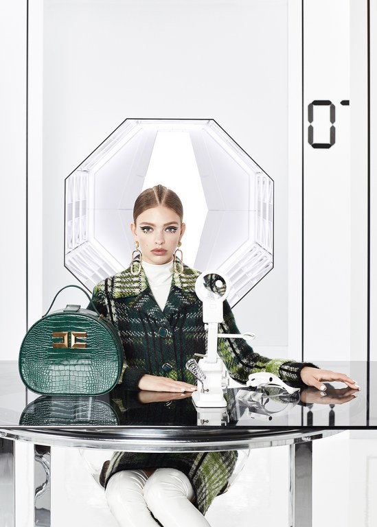 Fashionable adult woman wearing a plaid coat and white boots sitting at a table with a handbag and vintage telephone in a studio.