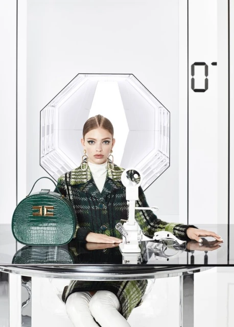 Fashionable adult woman wearing a plaid coat and white boots sitting at a table with a handbag and vintage telephone in a studio.