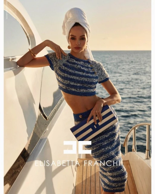 Fashion shot of a model on a yacht, wearing blue and white striped matching set with a white towel on her head and a clutch.