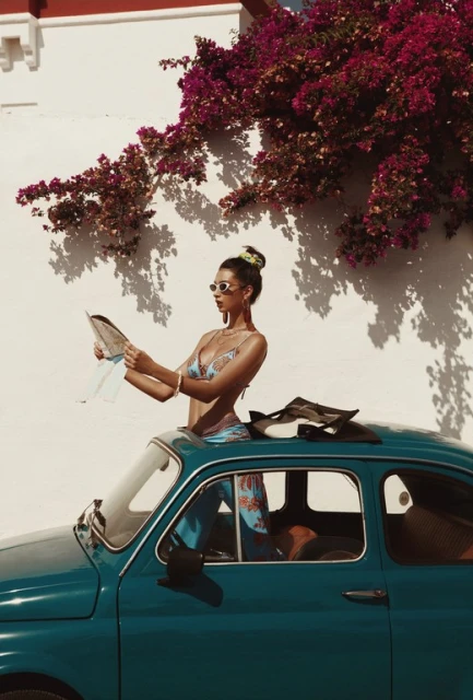 Woman in a swimsuit reading a map on top of a vintage teal car parked on a sunny street with bougainvillea on a white wall.