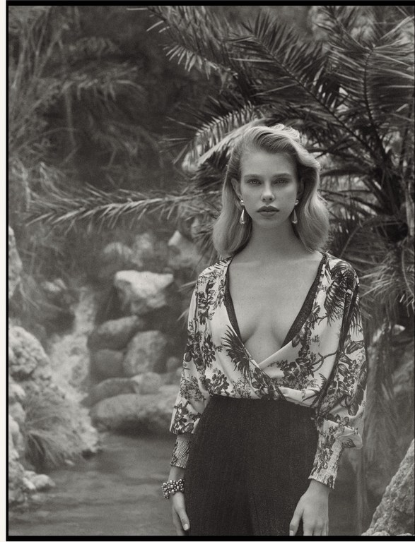 Monochrome portrait of a woman wearing a patterned V-neck top and dark trousers amidst tropical foliage and a rocky stream.