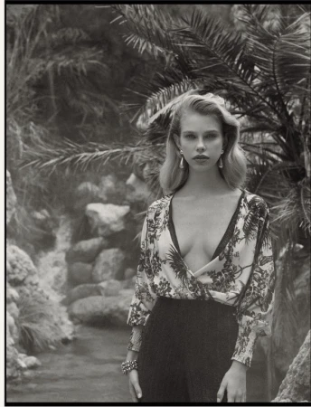 Monochrome portrait of a woman wearing a patterned V-neck top and dark trousers amidst tropical foliage and a rocky stream.