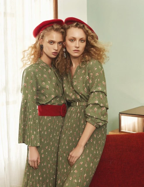 Two fair-skinned women with curly hair, wearing matching green floral print dresses and red berets, pose in an indoor setting.
