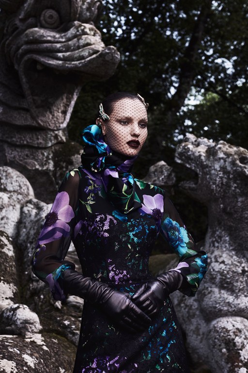 A woman in a floral dress and black gloves stands in a garden setting with stone sculptures and dark, shadowy lighting.