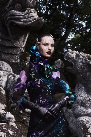 A woman in a floral dress and black gloves stands in a garden setting with stone sculptures and dark, shadowy lighting.