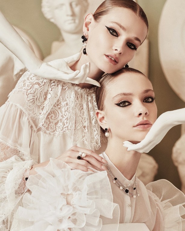 Two women in white dresses and jewelry pose in front of a statue; beauty, fashion, and art meet in a visually striking composition.