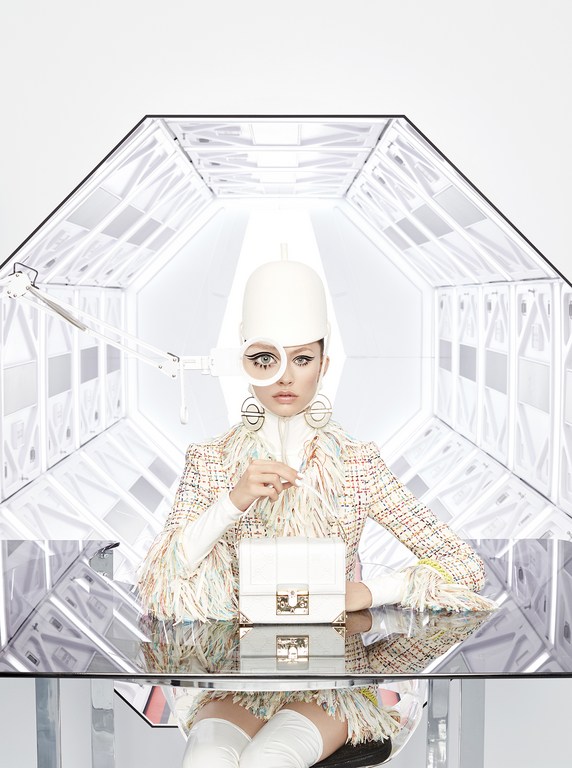 Fashion shot of a person wearing a tweed jacket and futuristic headwear, sitting at a reflective table in a white tunnel.