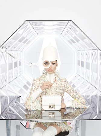 Fashion shot of a person wearing a tweed jacket and futuristic headwear, sitting at a reflective table in a white tunnel.