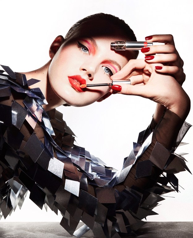 Beauty shot of a fair-skinned woman applying lipstick with makeup brushes in a photo studio, wearing a unique reflective clothing piece.