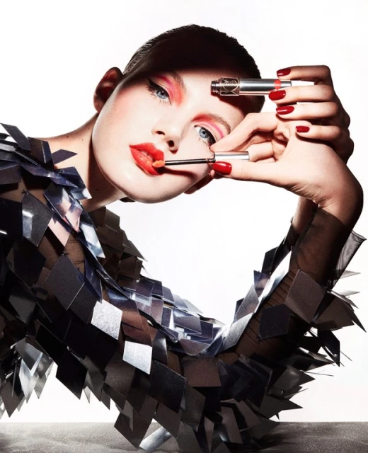 Beauty shot of a fair-skinned woman applying lipstick with makeup brushes in a photo studio, wearing a unique reflective clothing piece.